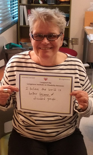 Catherine Kudlick, wearing thick-rimmed glasses has short grey hair and a big smile, while she holds her giving Tuesday #unselfie that says "I believe the world is a better place because of disabled people"